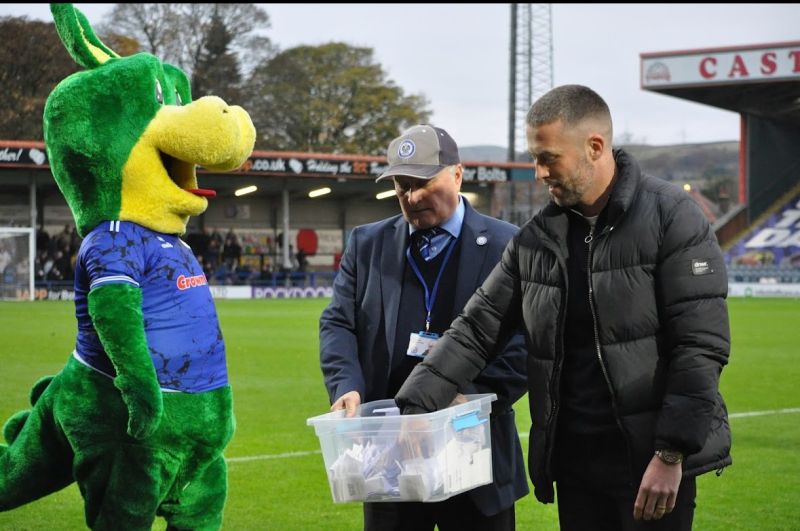 Tom Kennedy appearing as a guest at Rochdale's FA Cup match against Bromley. He is standing with the club's mascot on the pitch